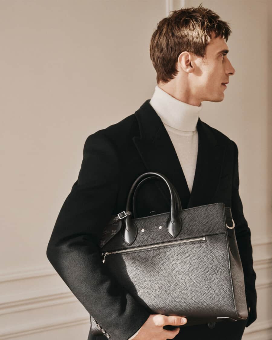 Man wearing white turtleneck and black overcoat carrying a luxury black leather Louis Vuitton tote bag under his arm
