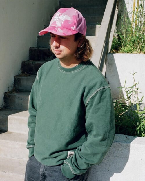 Man wearing loose green Stussy sweatshirt and pink baseball cap