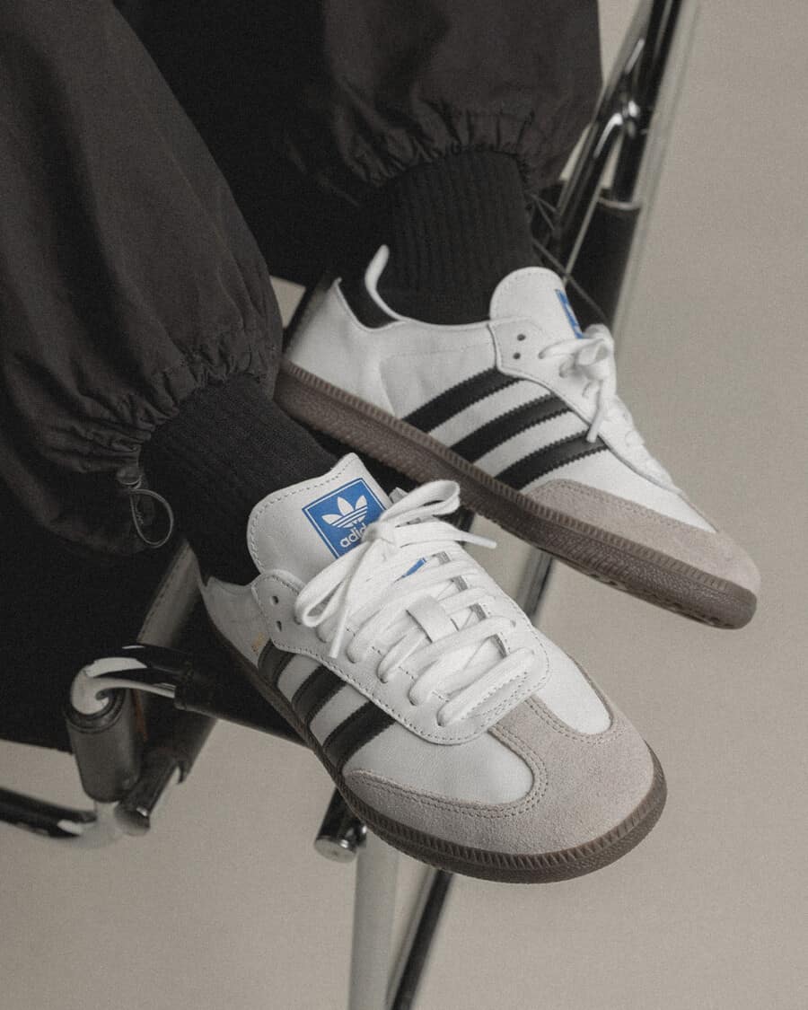 A pair of white affordable Adidas Samba sneakers worn on feet with black socks and pants