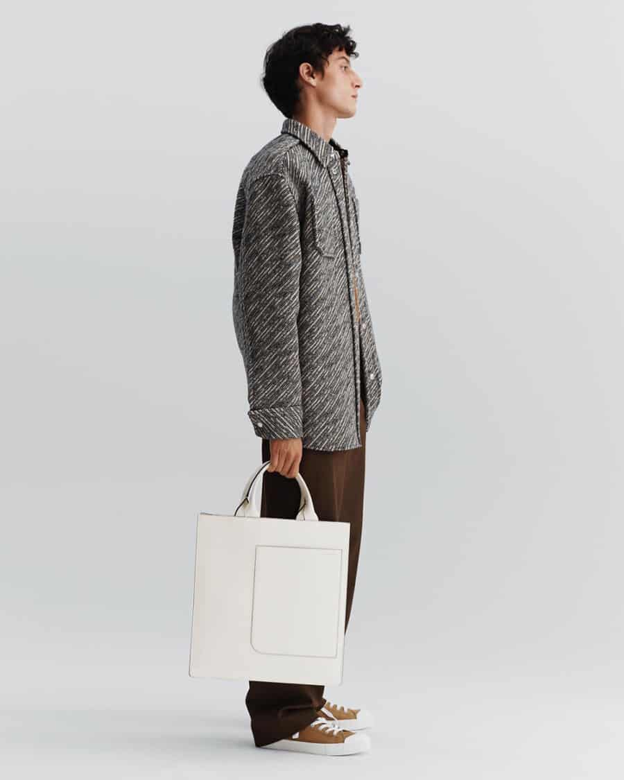 Man wearing loose brown pants, brown canvas sneakers and grey overshirt holding a luxury white tote bag