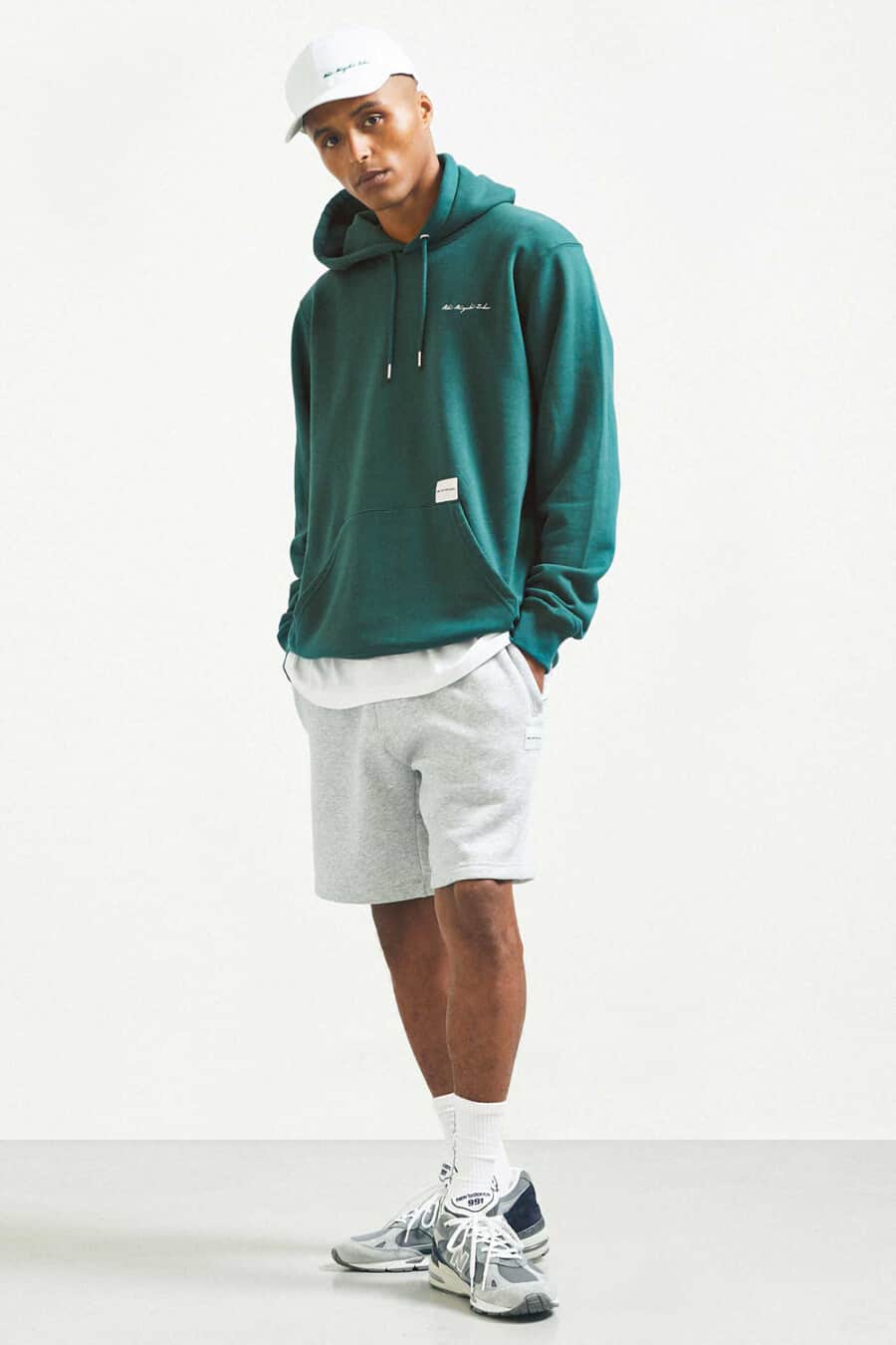 Men's grey jersey shorts, white T-shirt, green streetwear hoodie, white baseball cap, white tube socks and grey New Balance sneakers outfit