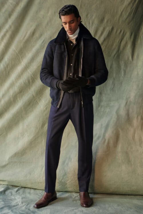 Men's luxury pleated navy trousers, white turtleneck, black shirt, navy shearling jacket and brown leather Chelsea boots outfit
