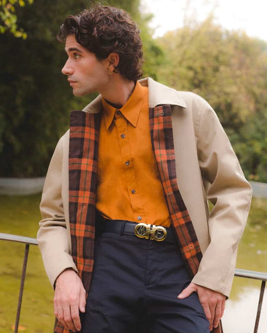 Man wearing navy pants, orange shirt, stone mac coat and a luxury branded Salvatore Ferragamo belt