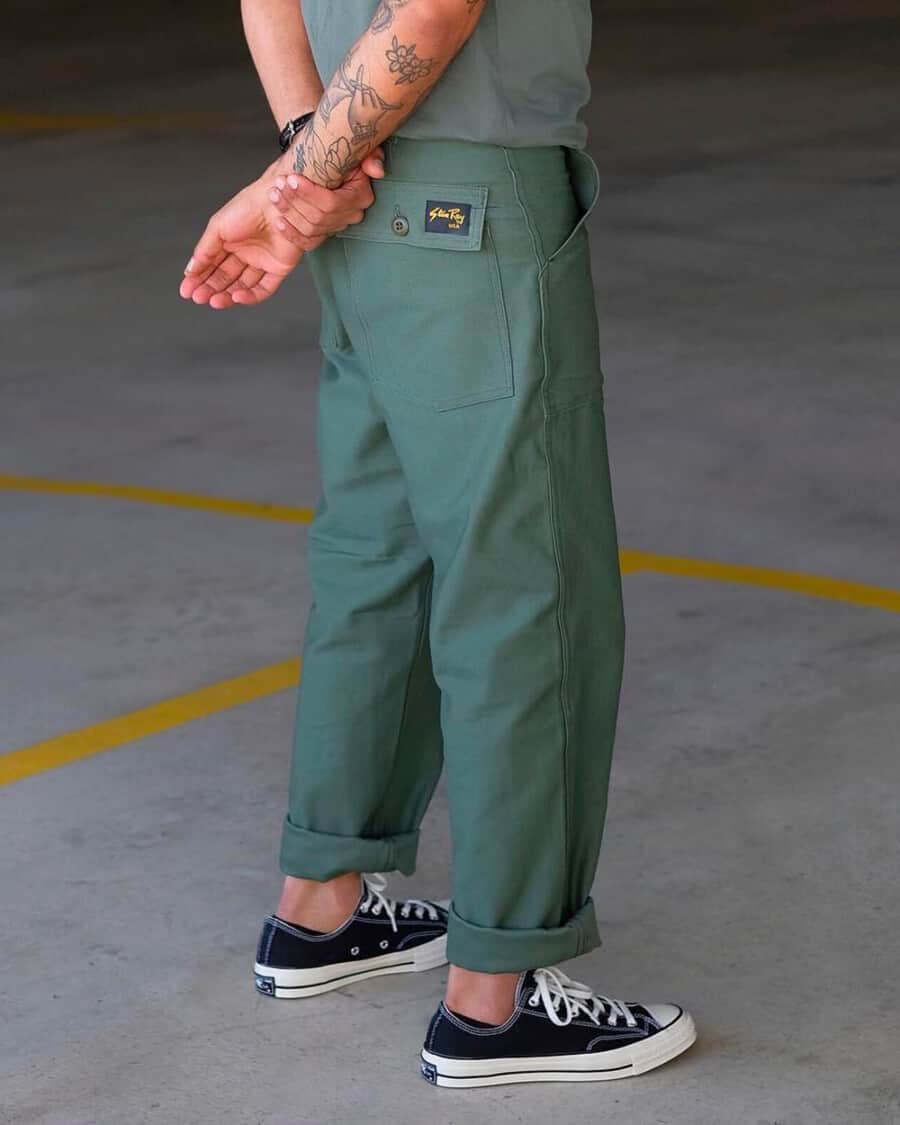 Man wearing green wide leg twill chino pants by Stan Ray with no socks and low-top Converse canvas sneakers in black