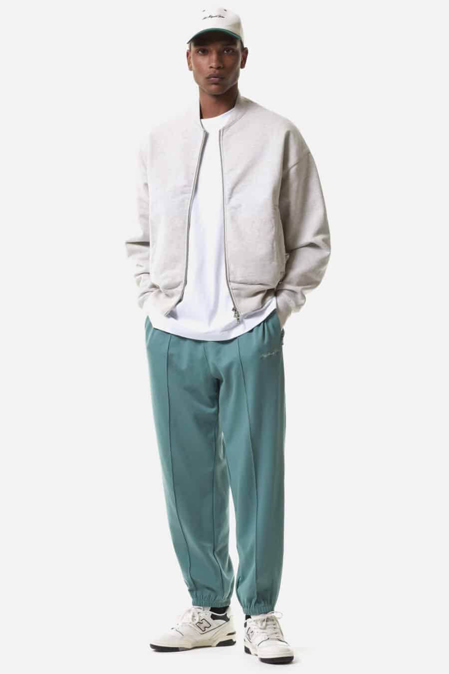 Men's green track pants, white T-shirt, stone bomber jacket, white baseball cap and white New Balance sneakers outfit