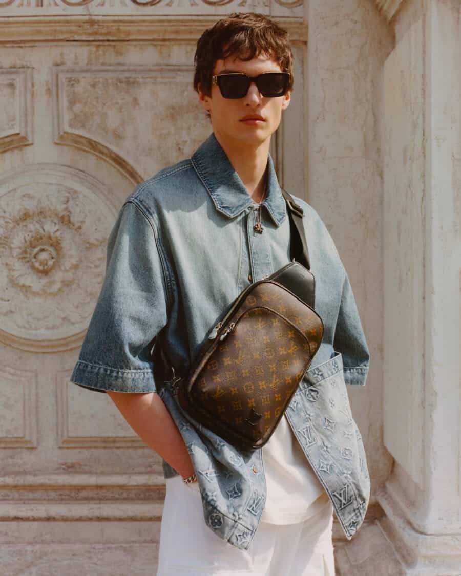 Man wearing a denim overshirt, white T-shirt and white pants with a luxury brown monogrammed Louis Vuitton crossbody bag