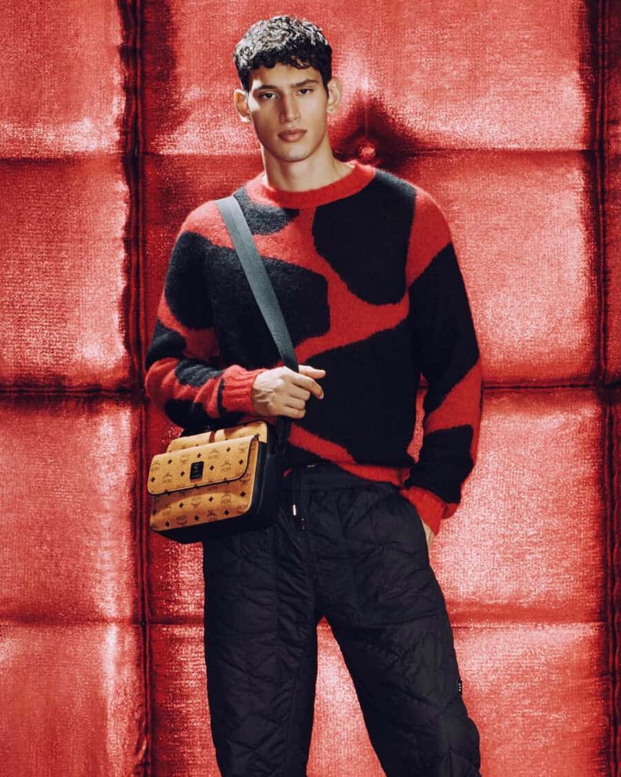 Man wearing black pants, red/black jumper and an MCM tan leather crossbody belt bag