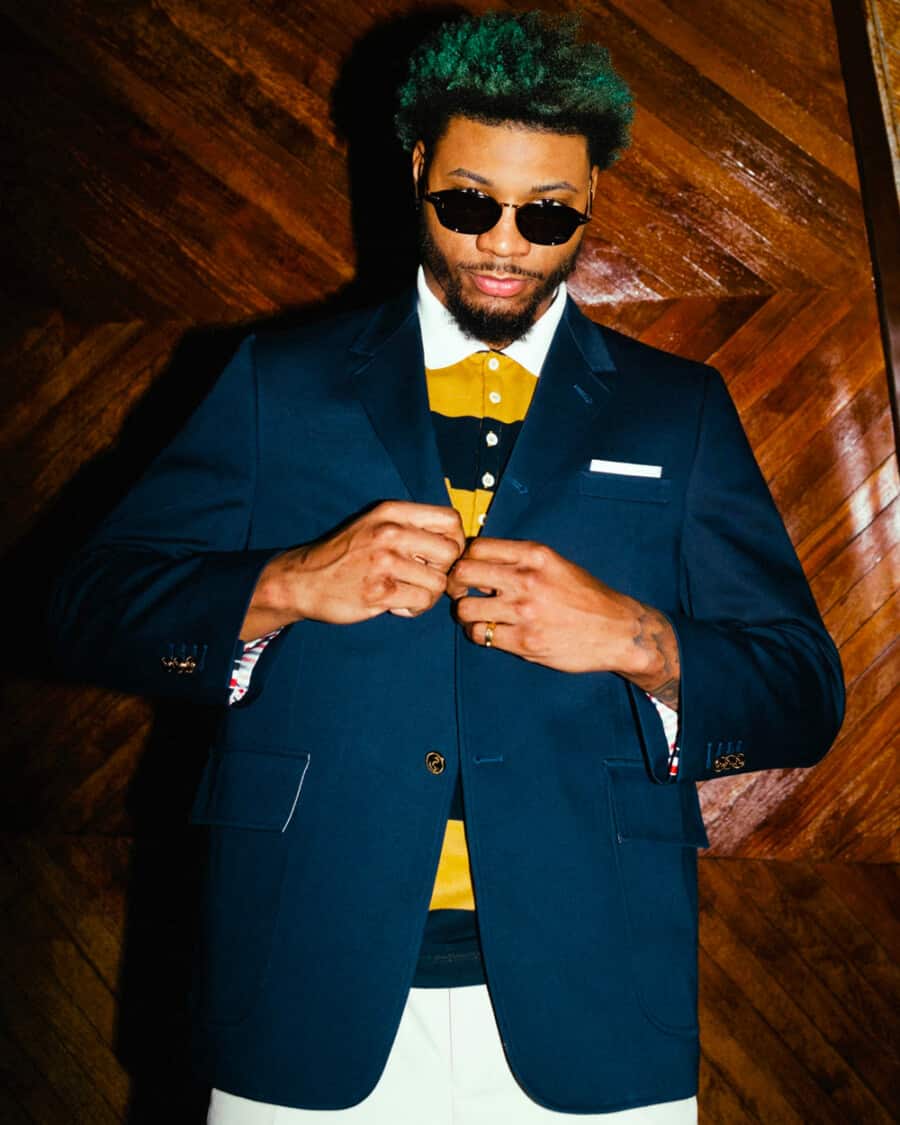 Black man wearing a mustard and navy striped Thom Browne polo shirt underneath a navy blazer with white pants