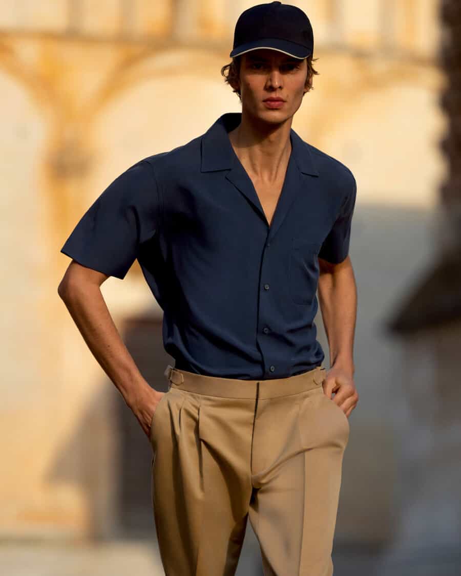 Man wearing high quality khaki pleated pants by Loro Piana with a navy Cuban collar short sleeve shirt and navy baseball cap