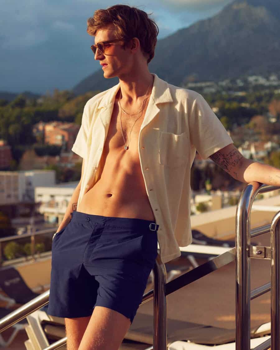 Man wearing navy tailored swim shorts with an open Cuban collar white shirt and sunglasses
