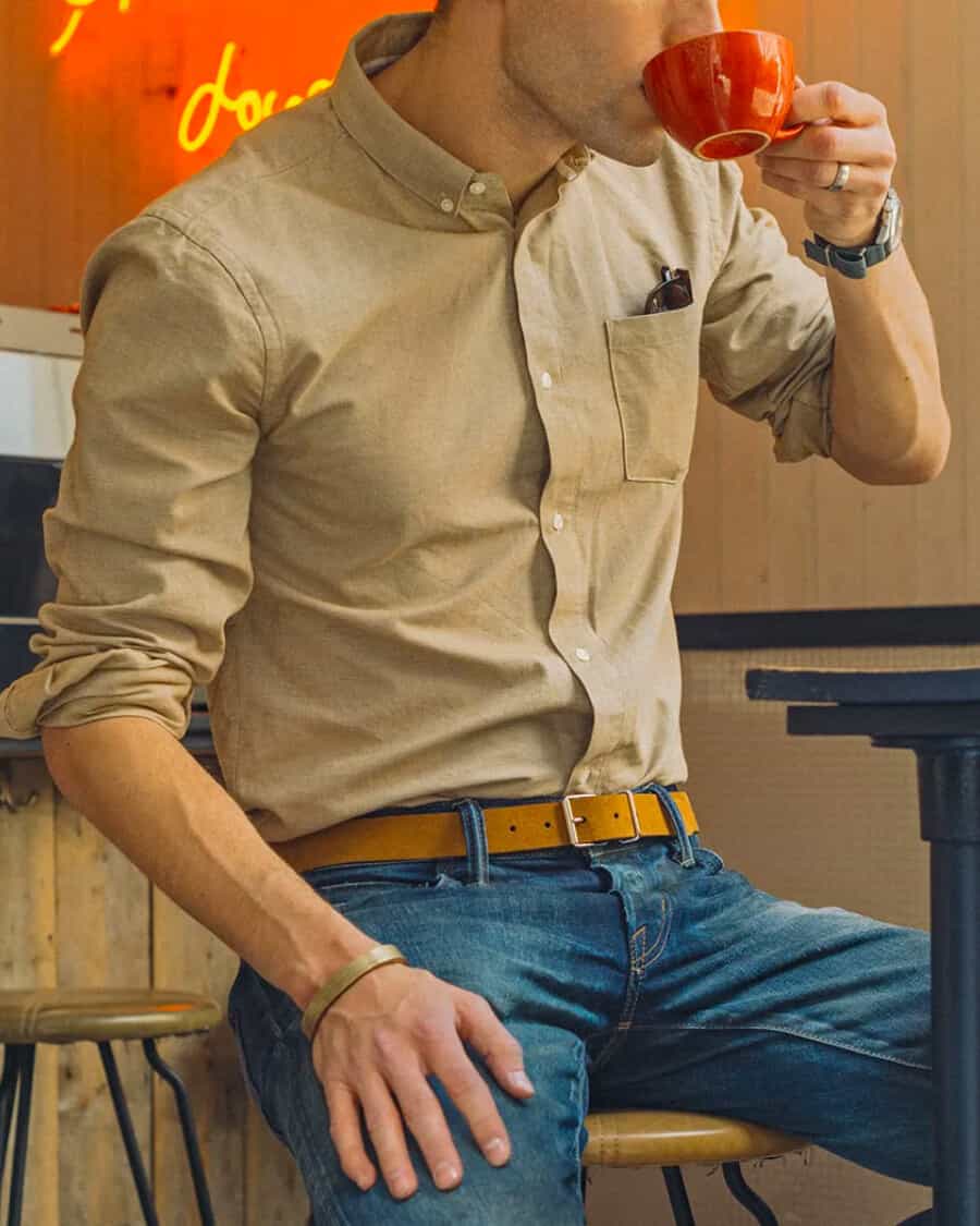 Man sitting drinking coffee wearing a khaki shirt, blue jeans and luxury tan suede belt