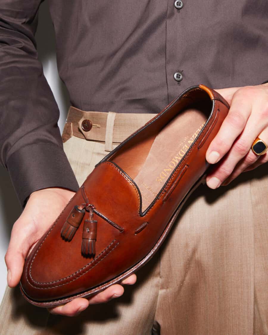 Man holding luxury brown leather tassel loafer while wearing khaki pants and black shirt