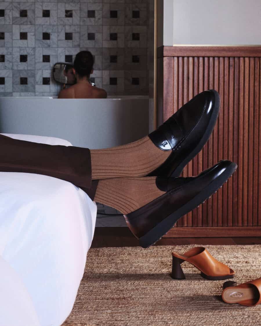 Man wearing a pair of Tod's shoes lying on a bed while woman has bath in background