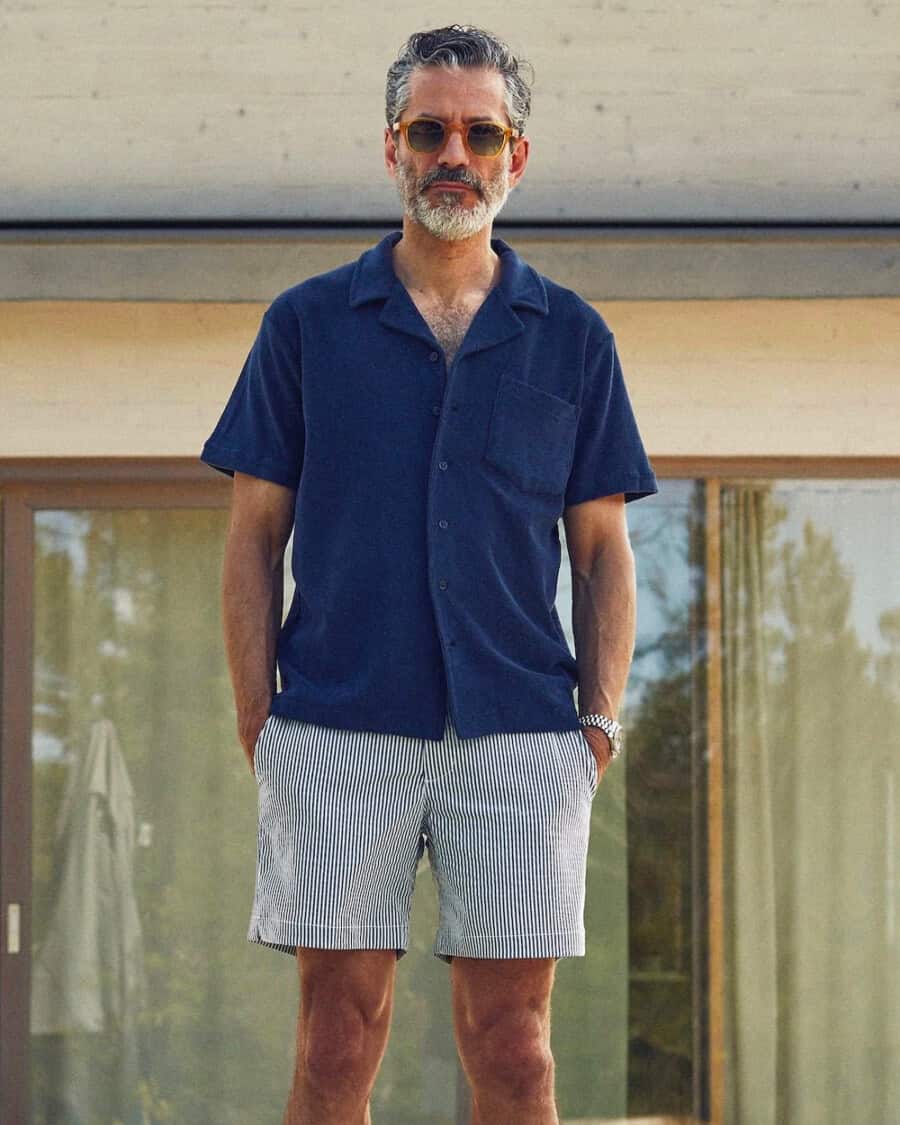 Man wearing striped seersucker swim shorts with a navy terry towelling polo shirt and sunglasses
