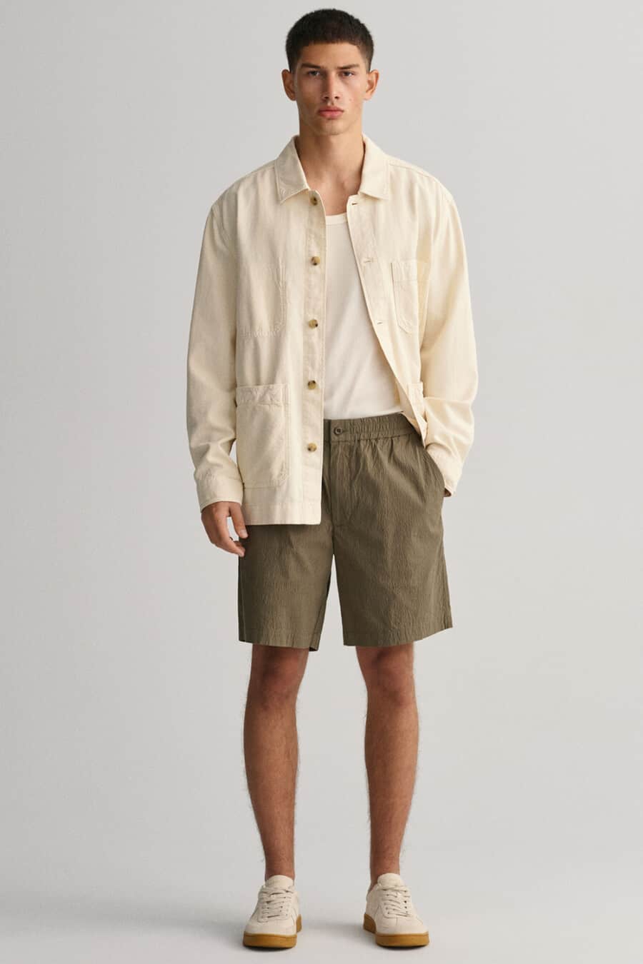 Men's olive green bermuda shorts, white vest, beige overshirt and off-white gum sole sneakers outfit