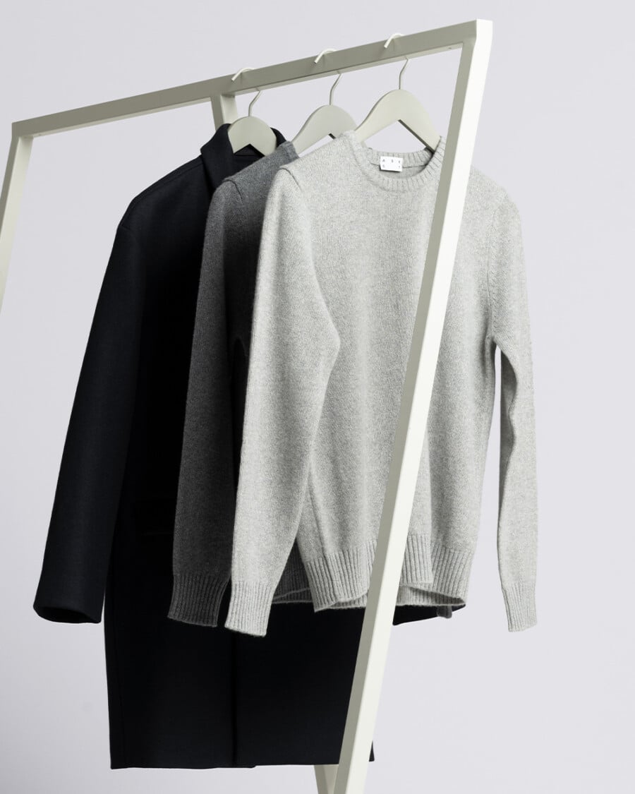 A rail of ASKET clothes including grey knitted sweaters and a black overcoat