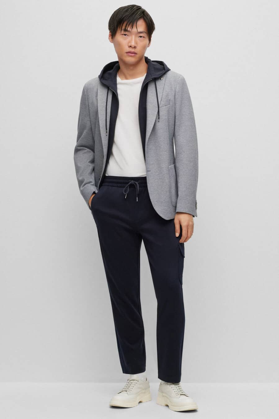 Men's navy drawstring pants, white T-shirt, navy technical hoodie, grey blazer and off-white sneakers outfit