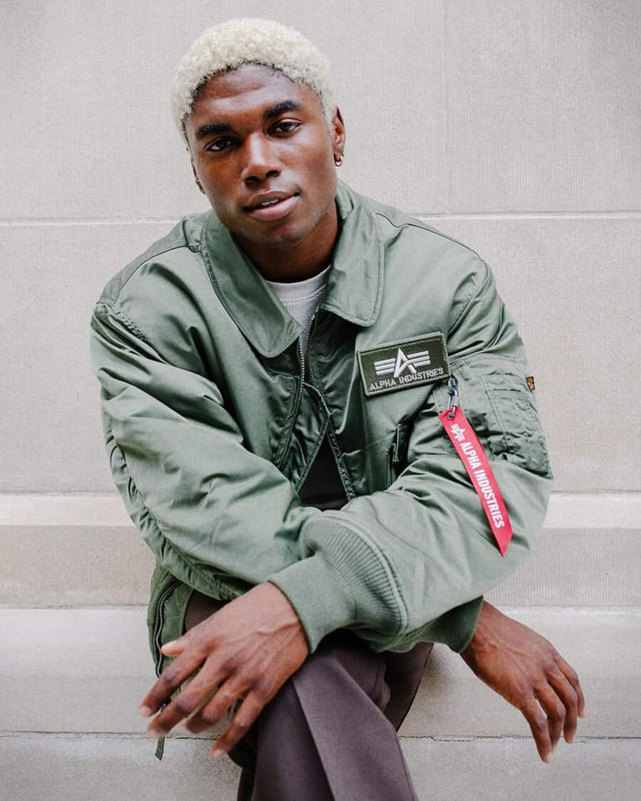 Black man with dyed hair wearing an Alpha Industries green MA-1 flight bomber jacket and brown pants