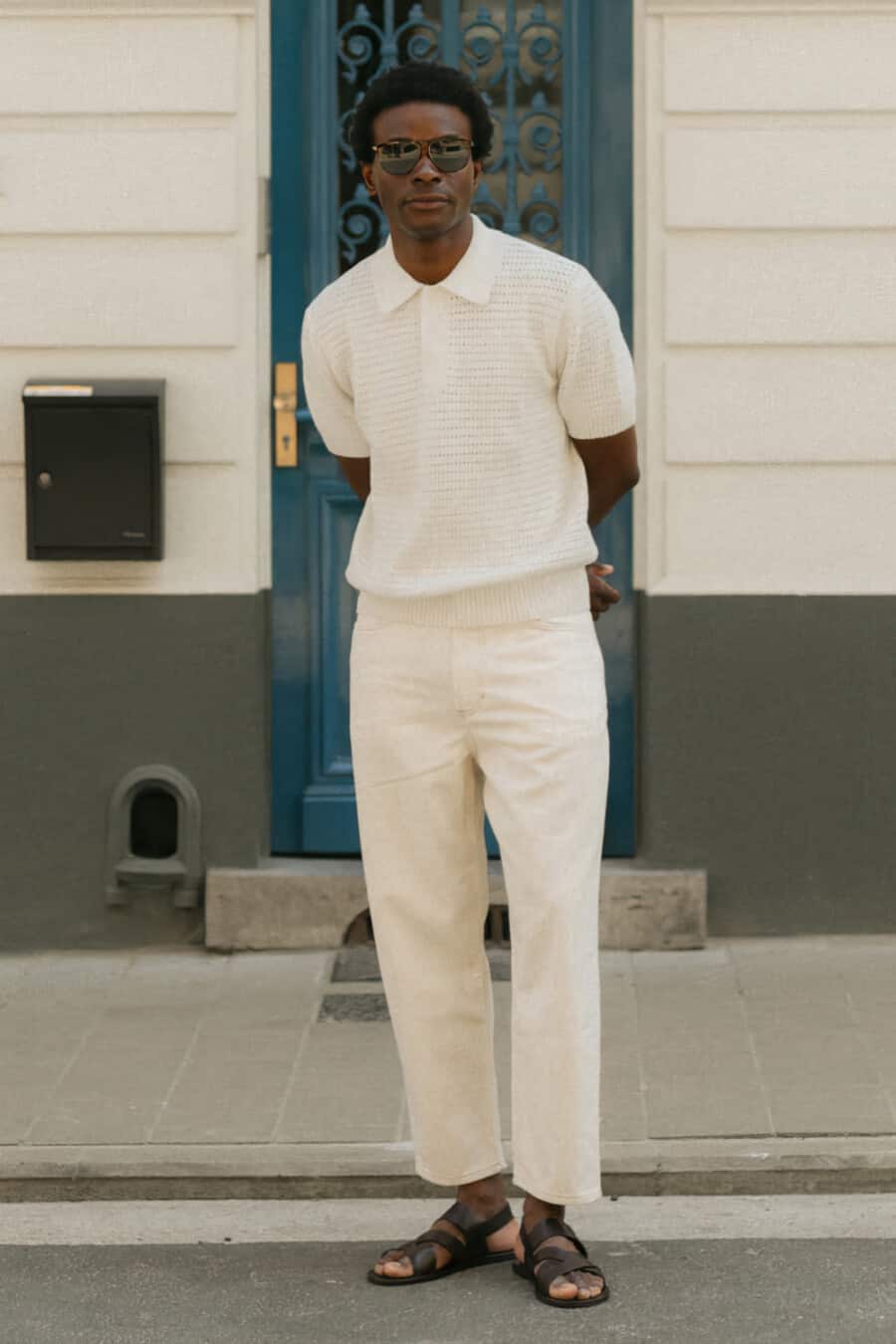 Black man wearing Mango knitted cream polo shirt, loose cream pants, brown leather sandals and tortoiseshell sunglasses