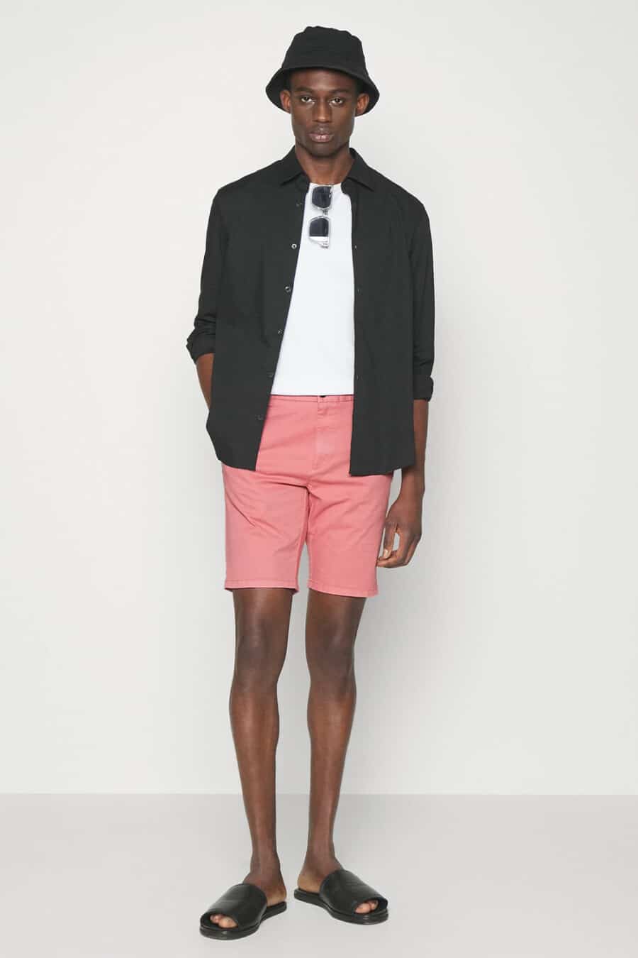 Men's bright pink shorts, white T-shirt, black open shirt, black bucket hat and black leather sliders outfit
