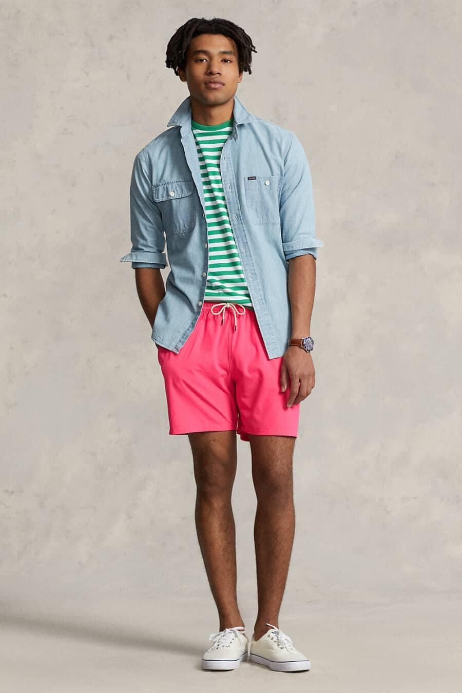 Men's bright pink swim shorts, white/green striped T-shirt, pale blue denim shirt and off-white canvas sneakers outfit