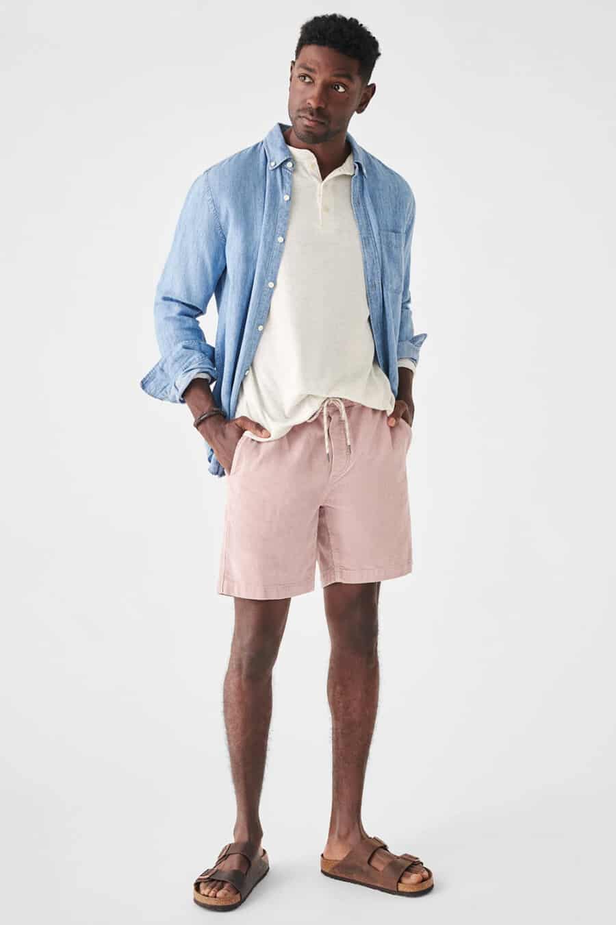 Men's light pink drawstring shorts, white Henley top, light blue linen shirt and brown leather slider sandals outfit