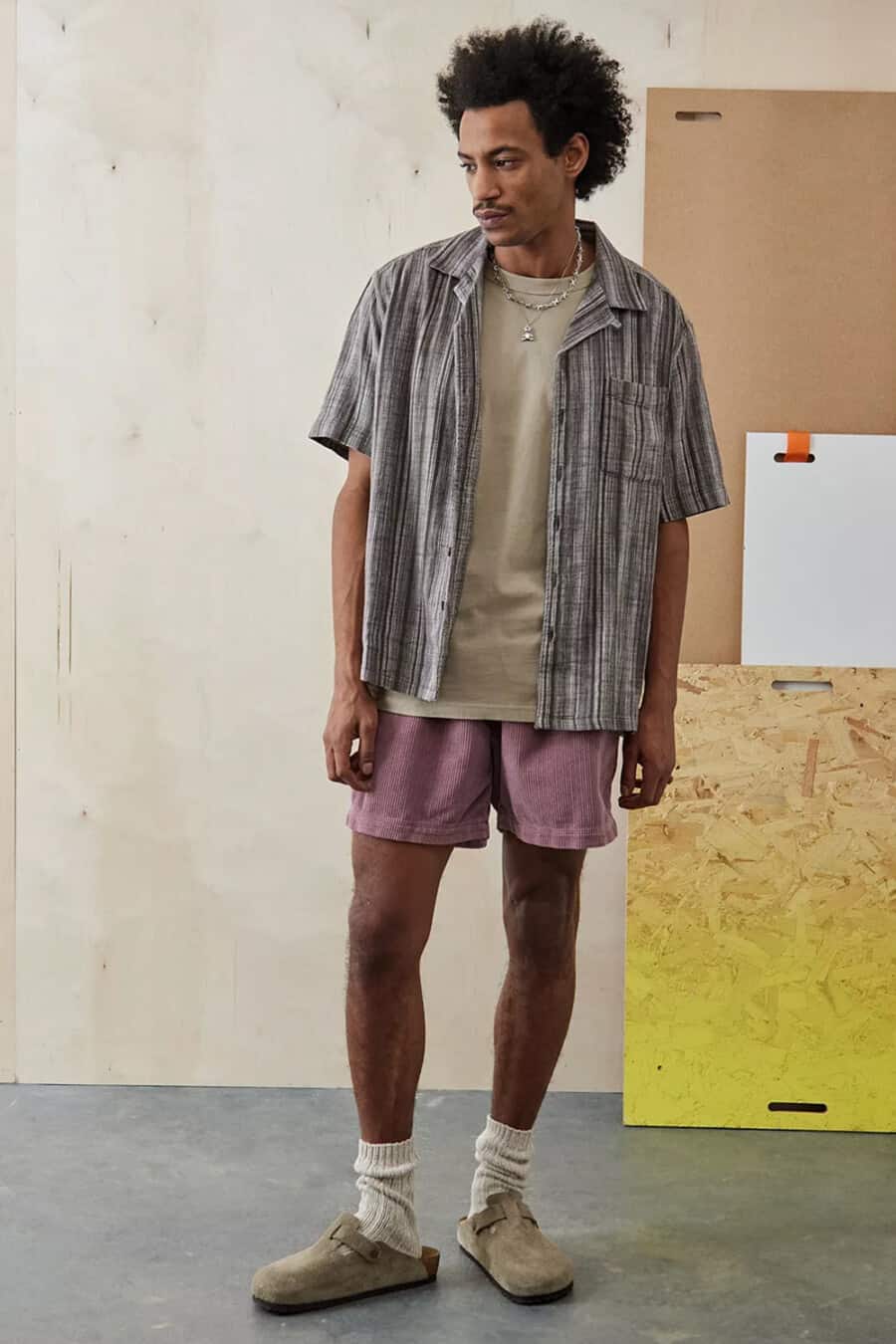Men's pink shorts, clay grey T-shirt, charcoal striped open short sleeve shirt, off-white socks and taupe suede mules outfit