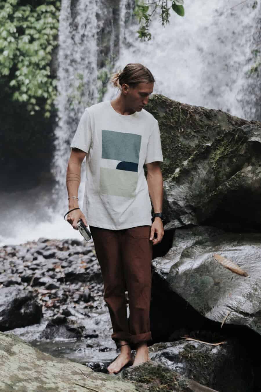 Man wearing brown pants, ethically made T-shirt in front of waterfall