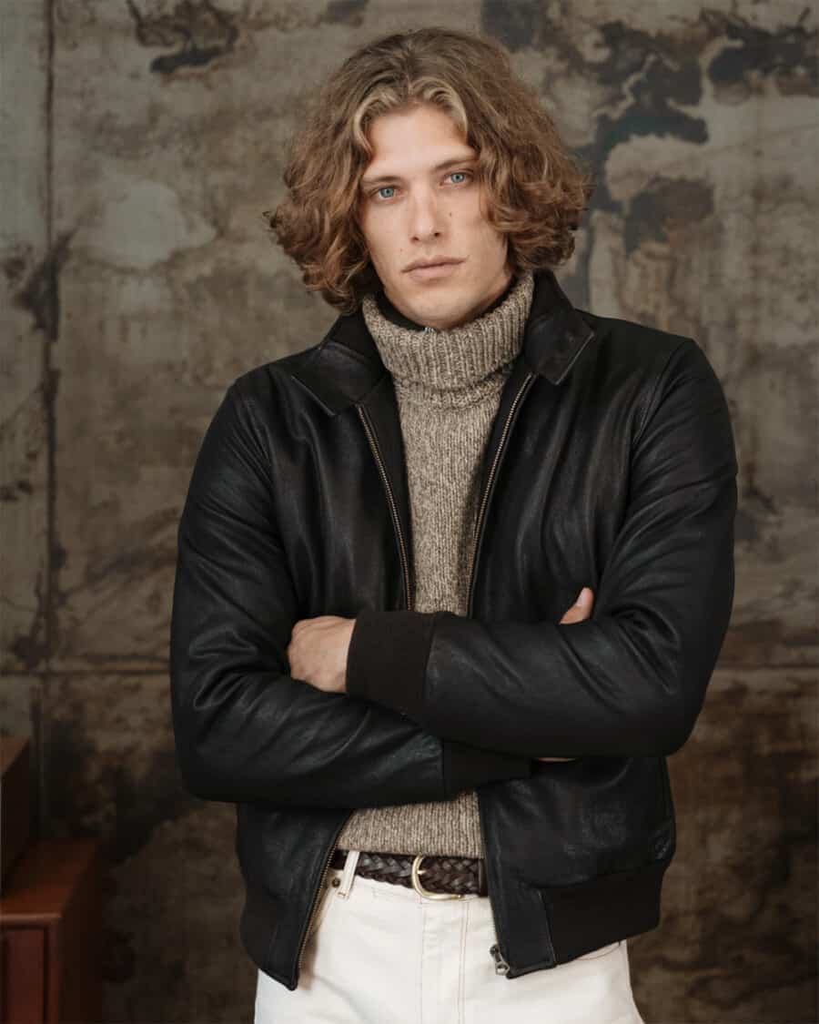 Man wearing a Velasca made in Italy black leather bomber jacket with brown chunky turtleneck and white pants