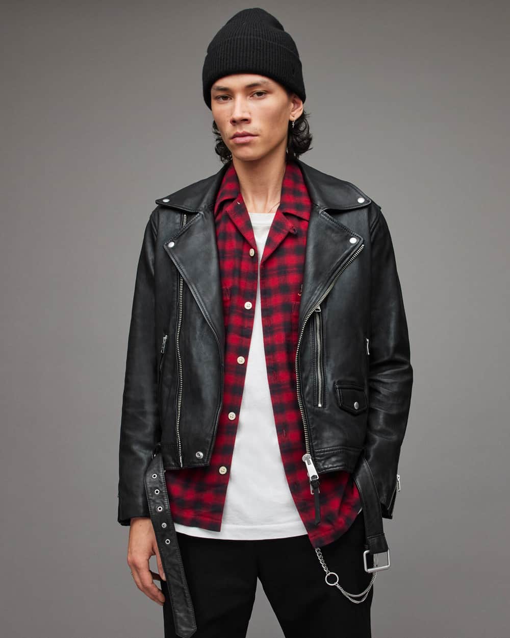 Man wearing ALLSAINTS black leather biker jacket with white longline T-shirt, red/black flannel shirt, black pants, black beanie and wallet chain