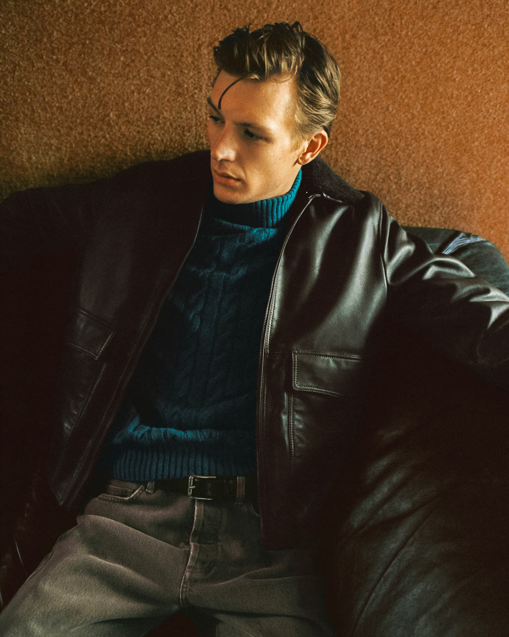 Man wearing black leather flight jacket with petrol blue cable knit turtleneck and grey jeans
