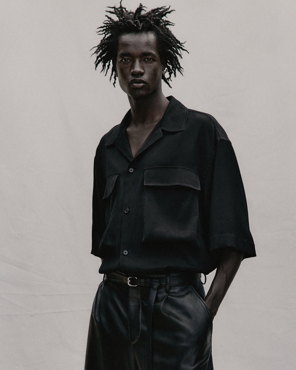 Black man wearing an oversized black short sleeve shirt and black faux leather pleated pants by Zara