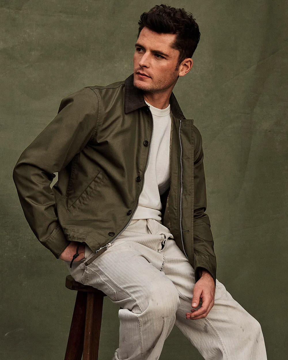 Man wearing stone pants by Buck Mason with an off-white sweatshirt and green collared twill jacket