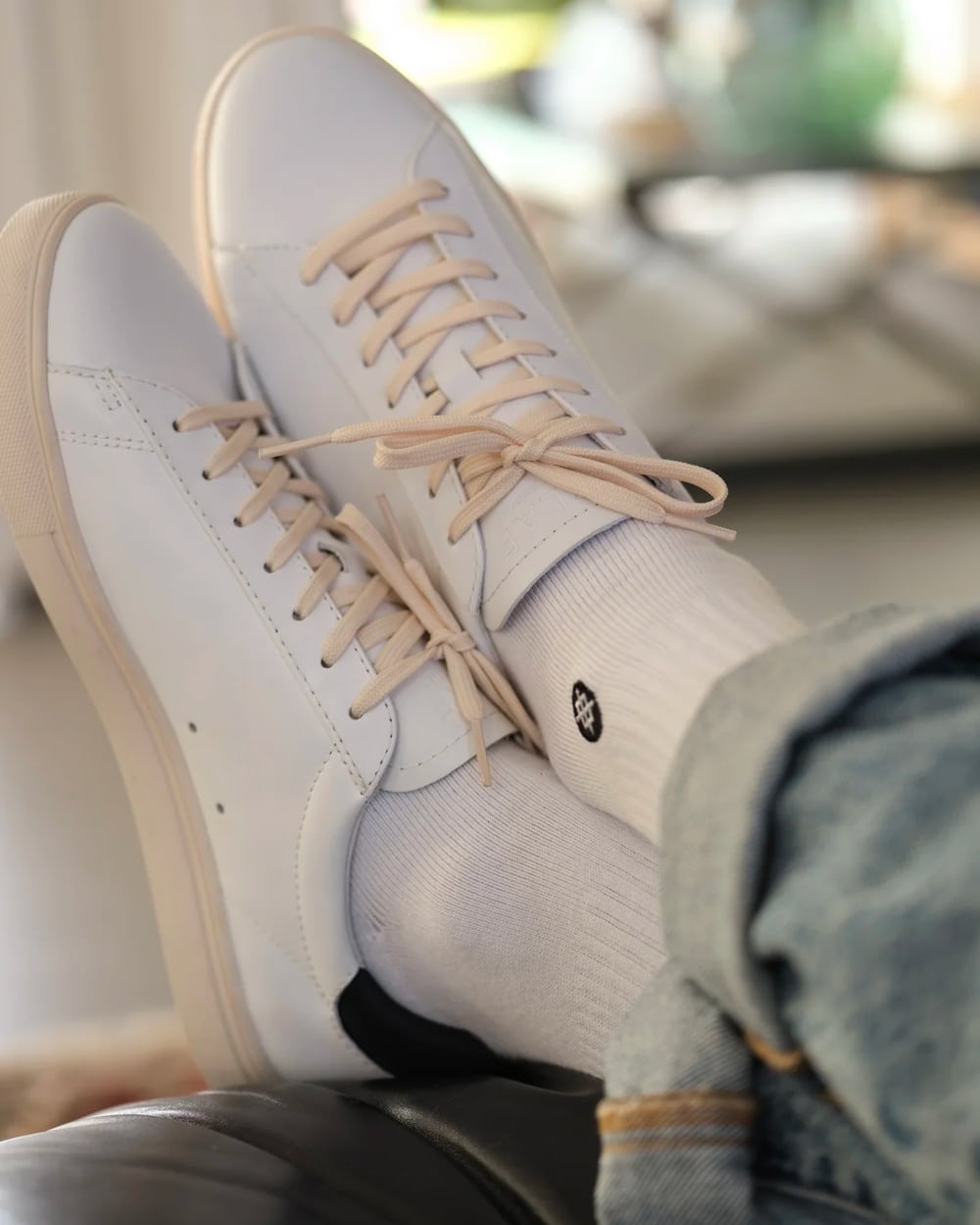 Pair of white leather CLAE sneakers with off-white outsole and laces worn on feet with white socks and light wash jeans