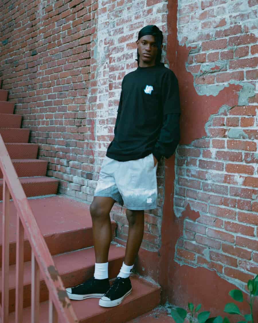 Black man wearing an Undefeated black sweatshirt, gradient blue shorts, white socks and black canvas sneakers