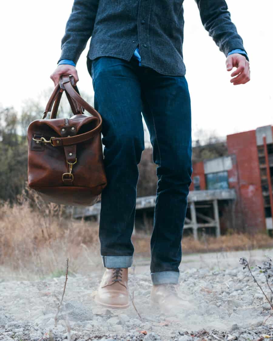 Man wearing Gustin selvedge denim jeans and grey flannel shirt holding a brown leather weekender bag