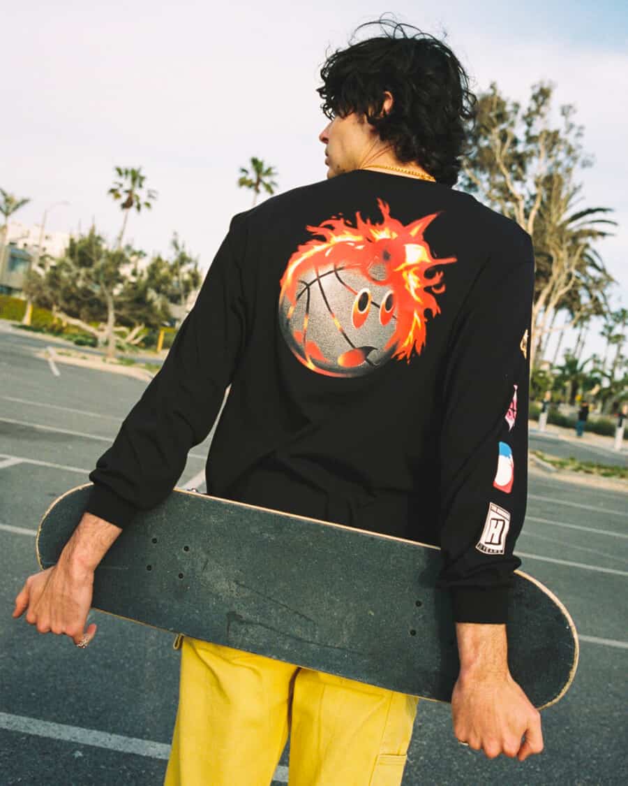 Skater wearing The Hundreds printed back black sweatshirt and yellow pants while holding a skateboard