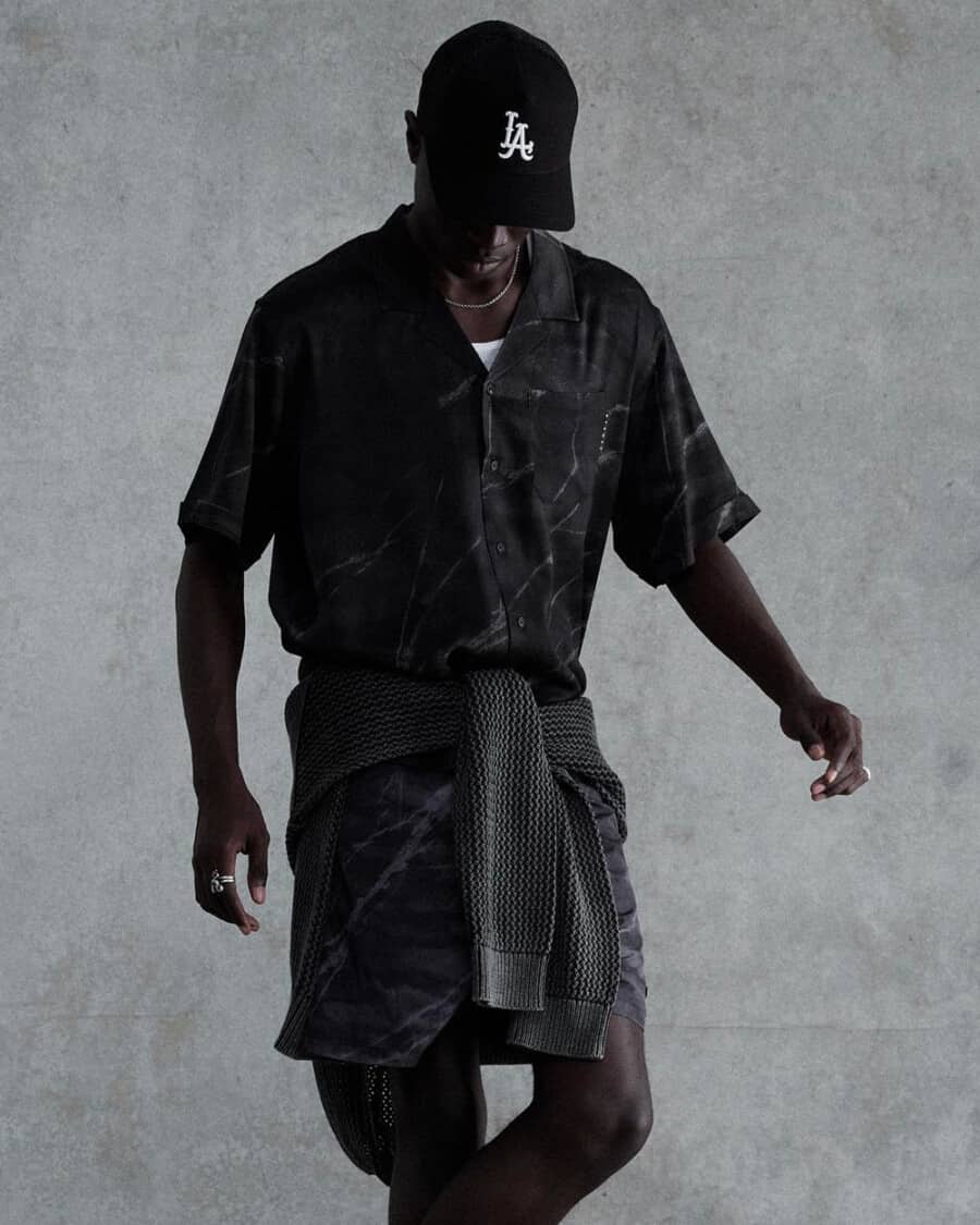 Man wearing black matching shirt and shorts, black LA baseball cap and tied grey sweater around the waist by Stampd