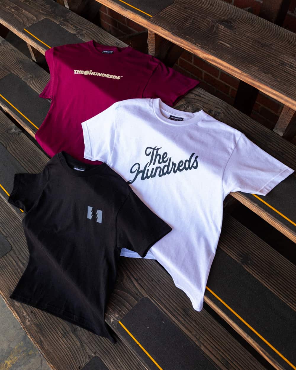 Three The Hundreds printed T-shirts in maroon, white and black laid out on bleachers