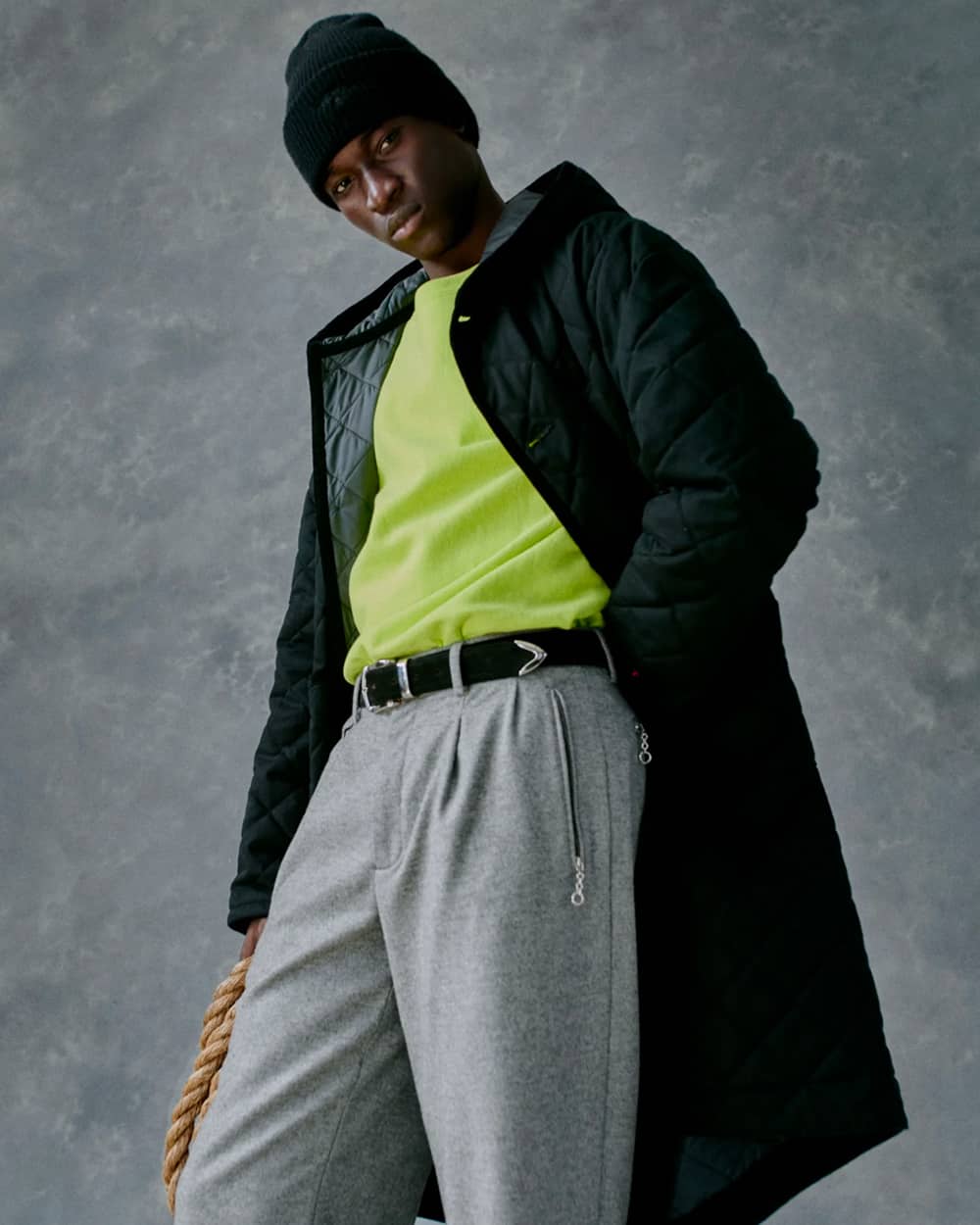 Black man wearing Noah NY bright green T-shirt, grey tailored sweatpants, long black down coat and green beanie