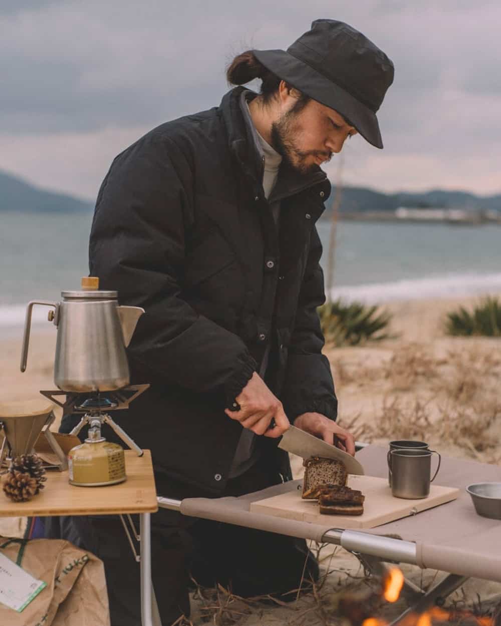 Man in the outdoors making tea and food wearing a Snow Peak black down jacket and black bucket hat