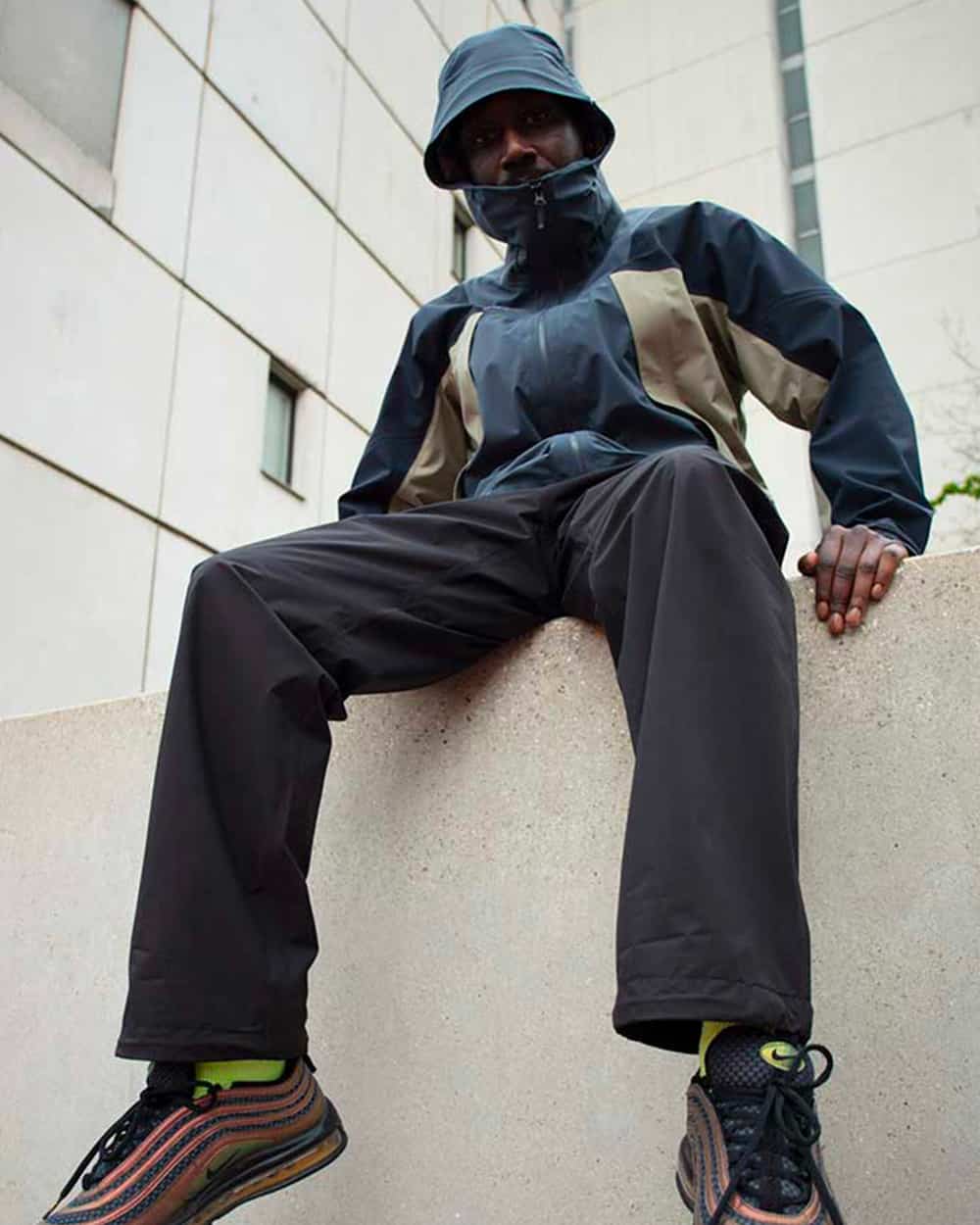 Man wearing a Goldwin technical wind breaker, blue bucket hat and charcoal work pants with multicoloured Nike sneakers