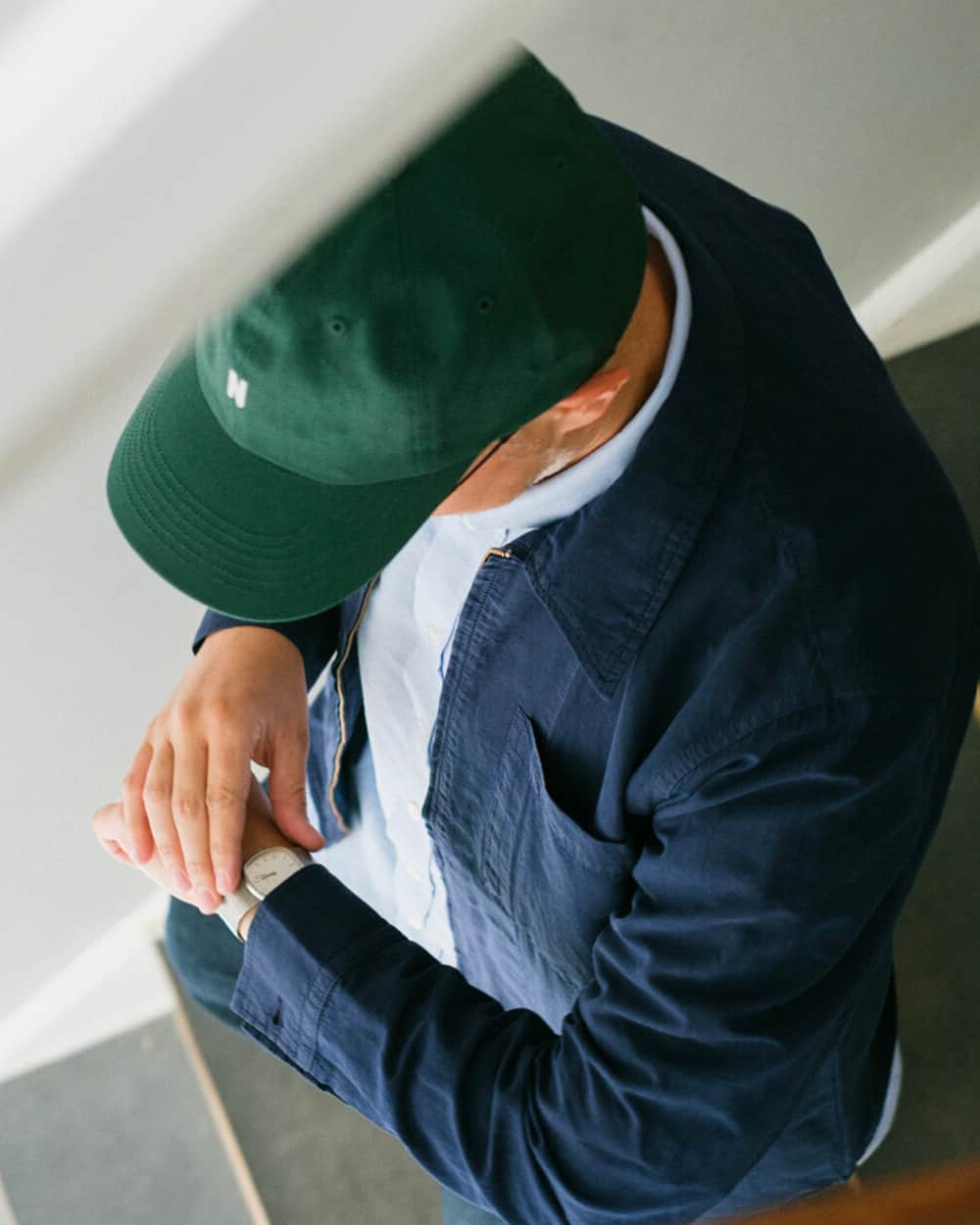 Man wearing a Norse Projects light blue T-shirt, dark blue collared lightweight jacket and green 'N' logo baseball cap looking at his watch