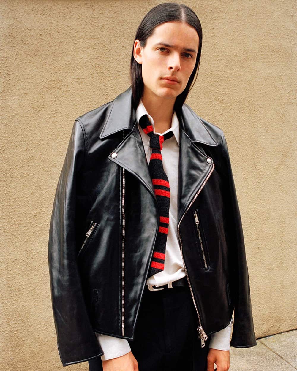 Man wearing an Our Legacy black leather biker jacket over a white shirt and black/red stripe tie with black pants