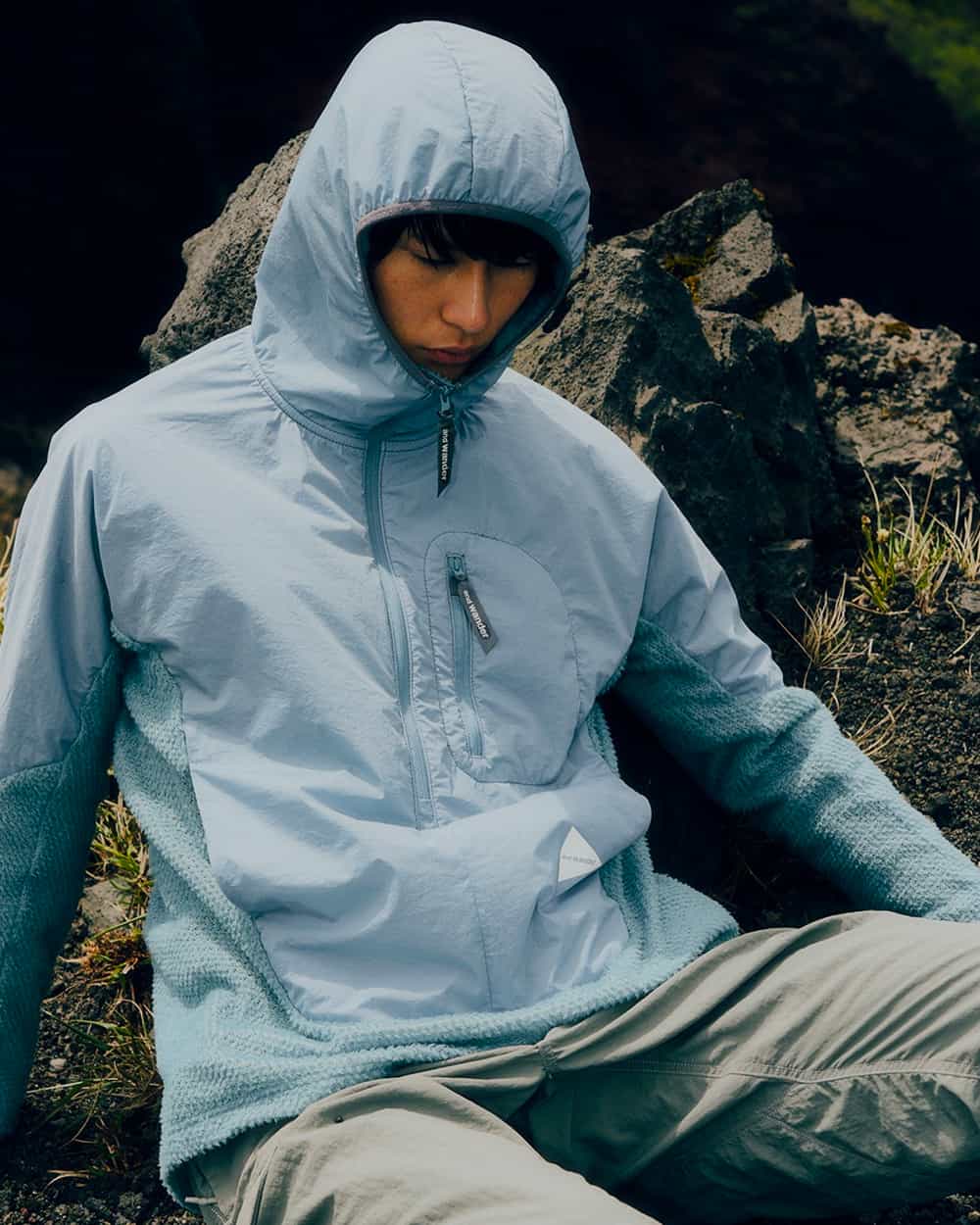Asian man wearing a light blue And Wander technical jacket with hood zipped up and grey technical pants