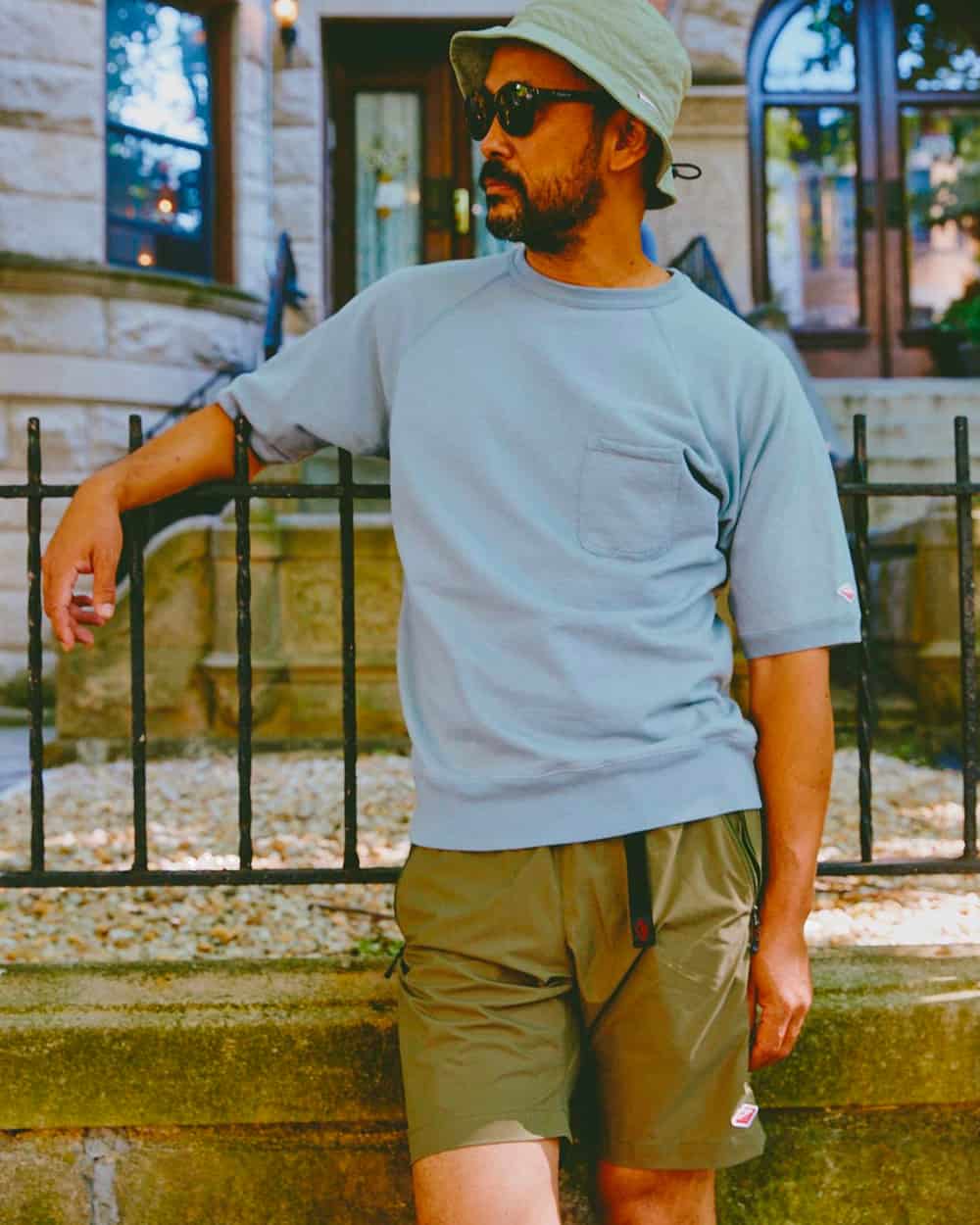 Man wearing a Battenwear light blue heavyweight T-shirt, khaki green athletic shorts, light green bucket hat and black sunglasses