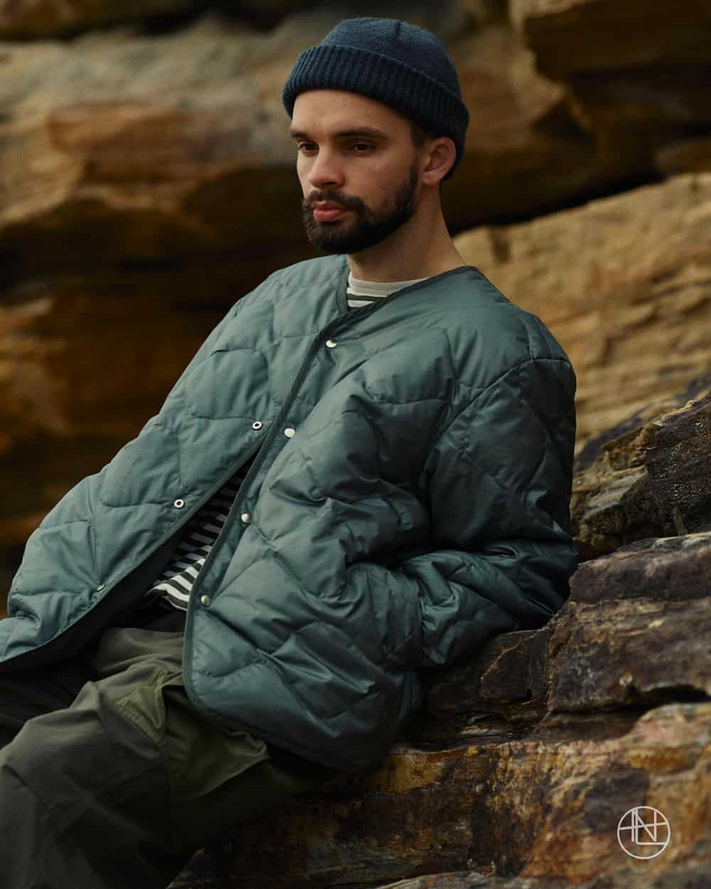 Man wearing a Nanamica quilted green-blue jacket, Breton stripe T-shirt, dark green pants and navy beanie