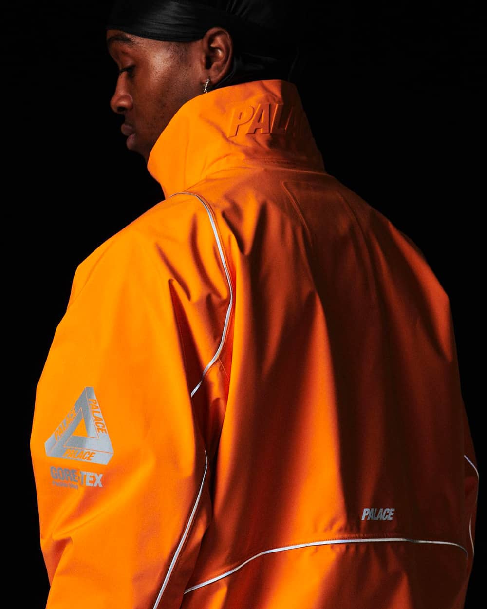 Black man wearing a bright orange Palace windbreaker jacket with logo on the arm