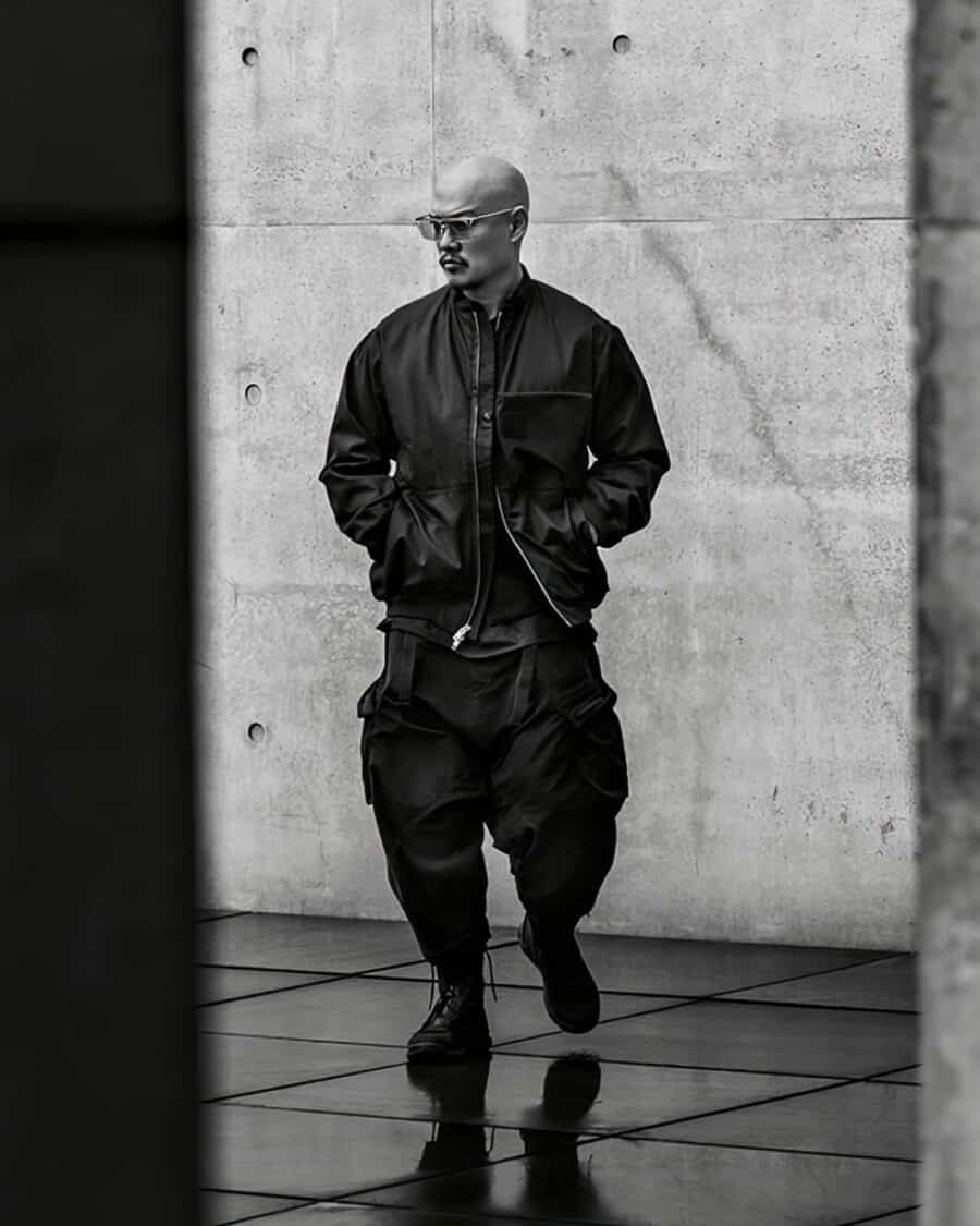 Man wearing Acronym men's all-black techwear, including a bomber jacket, loose combat pants and lace up boots