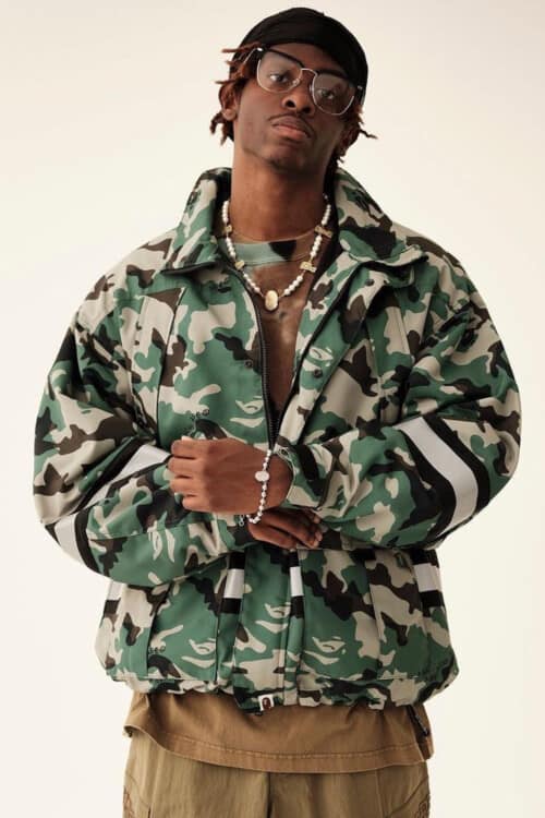 Black man wearing a camo A Bathing Ape / BAPE jacket over a brown T-shirt with khaki cargo shorts and eye glasses